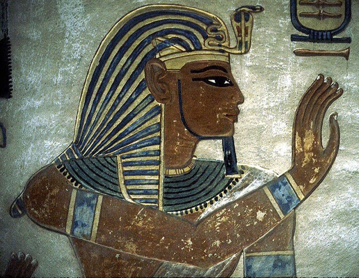 Egyptian Pharaoh dressed in blue and gold Egyptian dressage. He is holding his arm out to the left with his hand out with all five fingers. He is also facing to the right. 