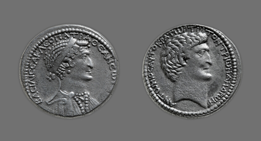 Double-sided coin. On the left, one side of the coin displays the head of Cleopatra VII facing the right and she dons a Macedonian diadem and a beaded necklace, her corkscrew curls bunned low. On the right, the other side of the coin depicts the head of Mark Antony as an exact male copy of Cleopatra VII.